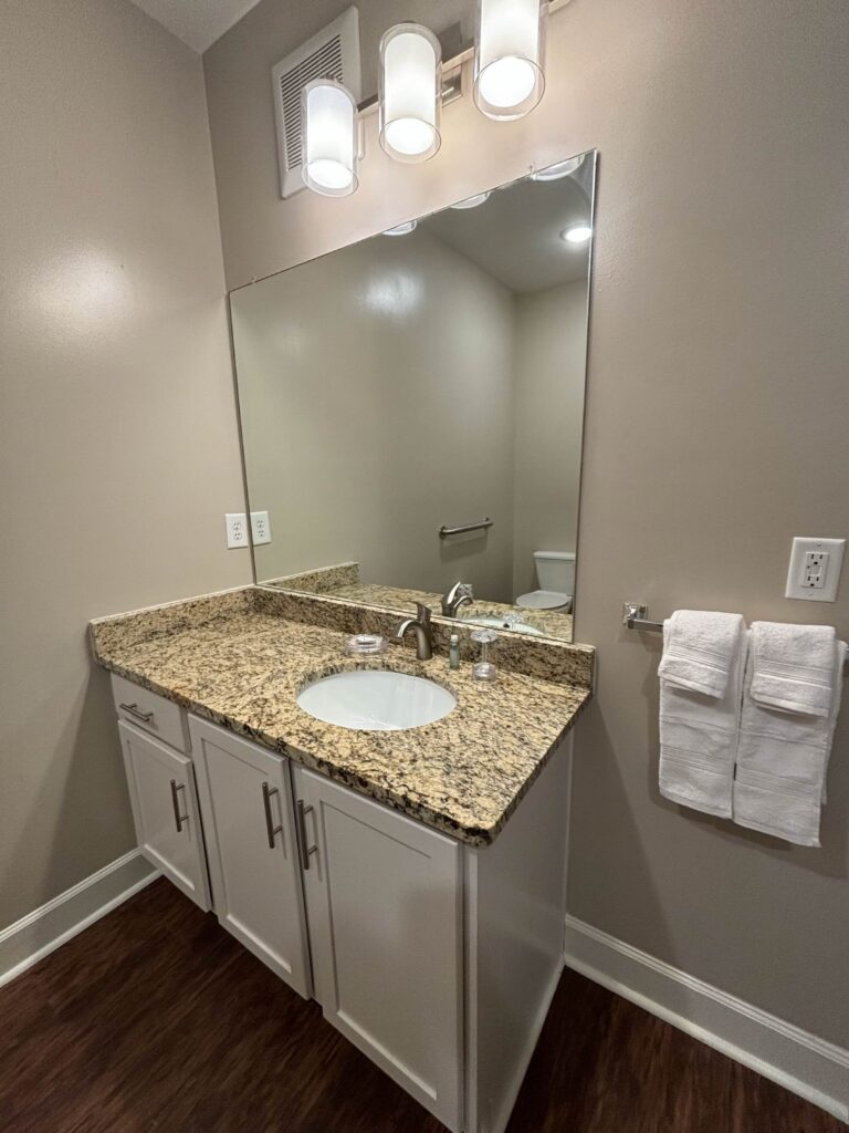Modern bathroom featuring a granite vanity countertop, white cabinetry, large wall mirror, contemporary lighting, and fresh white towels.