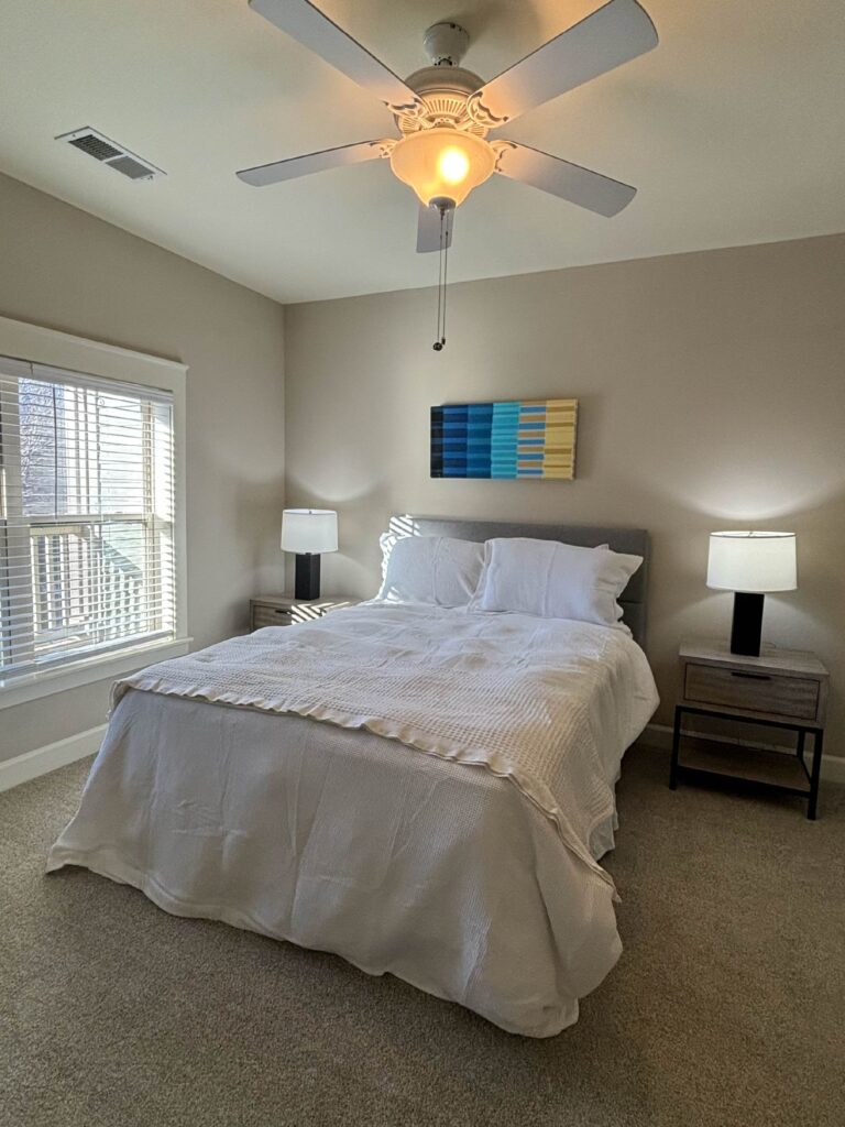 Primary bedroom at Weirbridge Village featuring a queen-size bed, matching nightstands with lamps, plush carpeting, ceiling fan, large window with natural light, and modern wall art.