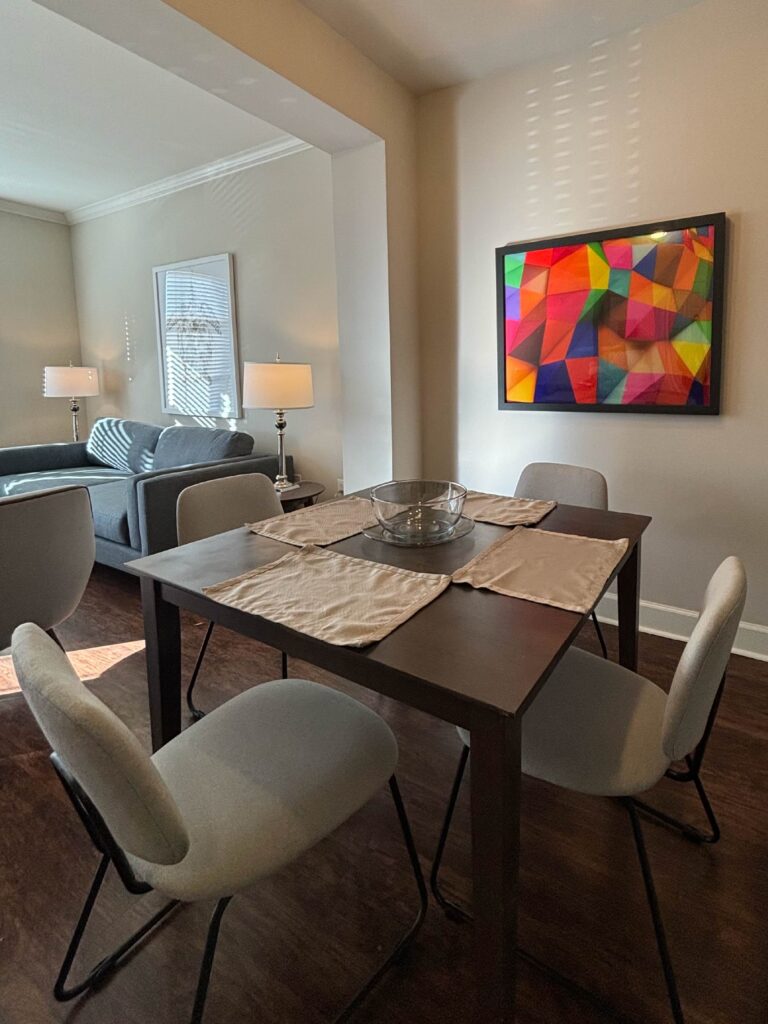 Modern dining area featuring a dark wood table with seating for four, upholstered chairs, contemporary artwork, and an open layout connecting to a bright living room.