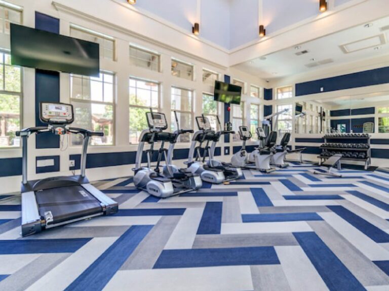 Modern fitness center at Weirbridge Village featuring treadmills, ellipticals, stationary bikes, free weights, strength training equipment, large windows, and high ceilings with abundant natural light.