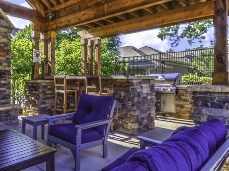 Covered outdoor pavilion at Weirbridge Village featuring cushioned lounge seating, stone accents, bar-height seating, built-in grilling station, and scenic community surroundings.