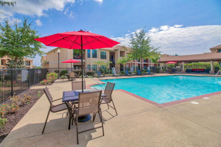Community swimming pool at Vista Ridge Apartments in Tupelo, Mississippi, featuring poolside seating, shaded tables with umbrellas, landscaped grounds, and a spacious sun deck.