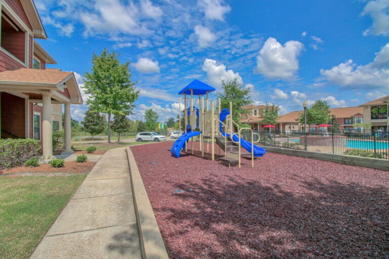 Outdoor playground area at Vista Ridge Apartments in Tupelo, Mississippi, featuring play structures, landscaped grounds, walking paths, and a nearby resort style swimming pool.