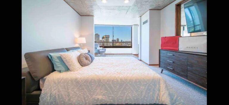 Spacious bedroom with a large bed, dresser, floor-to-ceiling window, and city views extending toward a private lanai.
