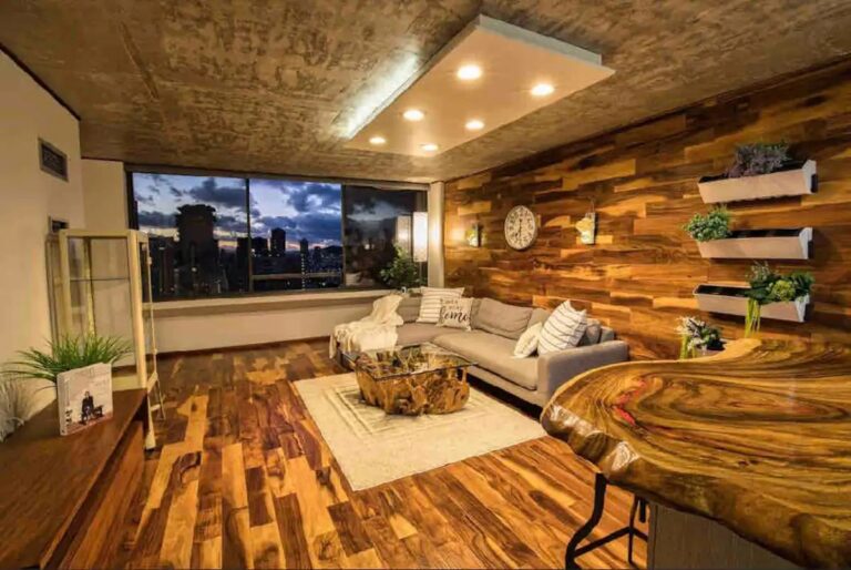 Warm, upscale living room with rich wood finishes, sectional sofa, custom wood accent walls, and large windows showcasing Waikiki skyline views.