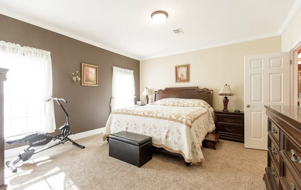Large primary bedroom featuring a king-size bed, plush carpeting, traditional wood furnishings, and a compact exercise machine near bright windows.