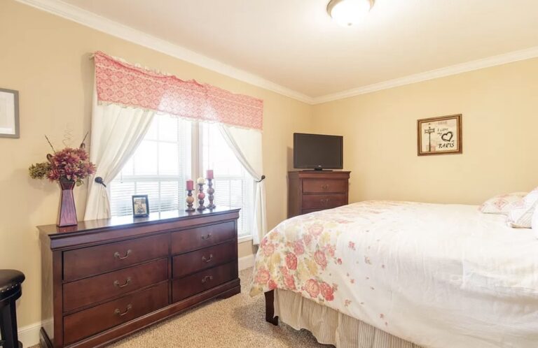 Comfortable guest bedroom featuring a queen bed, dark wood furnishings, soft carpeting, and natural light from a large window.
