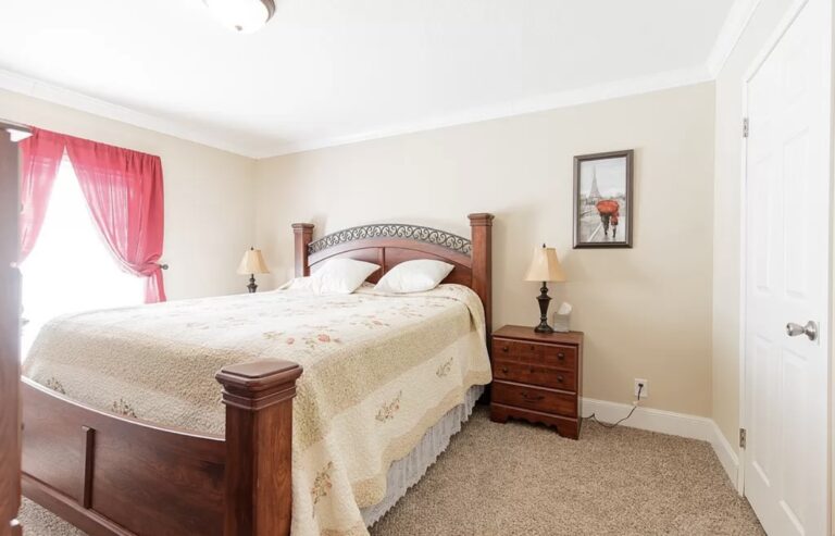 Spacious bedroom featuring a king-size bed with a decorative wood headboard, plush carpeting, bedside tables, and natural light from a large window.