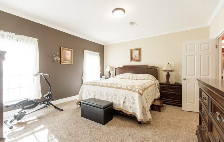 Large primary bedroom featuring a king-size bed, plush carpeting, traditional wood furnishings, and a compact exercise machine near bright windows.