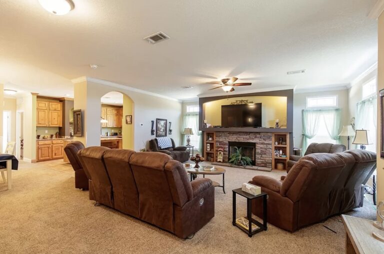 Open-concept great room with reclining sofas, stone fireplace, large television, and adjoining kitchen access.