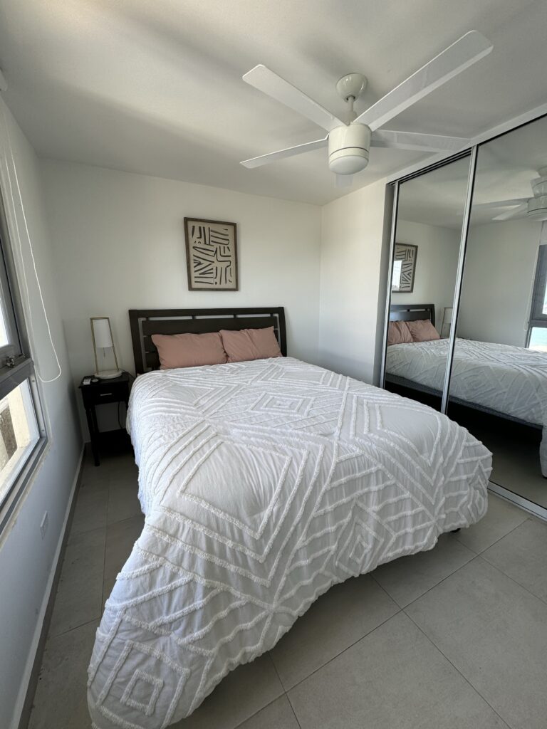 Modern bedroom with a queen bed, mirrored closet doors, ceiling fan, bedside table, and large window providing natural light.